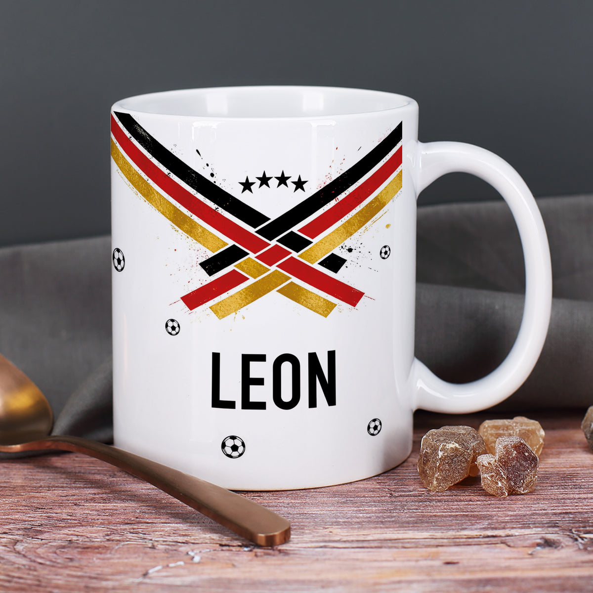 Personalisierte Fußball Tasse mit Name Leon, Fan-Design in Schwarz Rot Gold, kleine Fußbälle und Zuckerstücke im Hintergrund