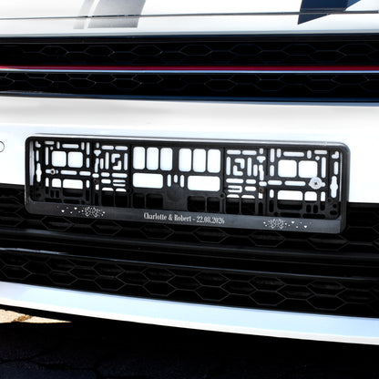 Schwarzer Nummernschildhalter am Auto mit Wunschtext Charlotte und Robert, Hochzeitsdatum und Tauben Herzen Motiv