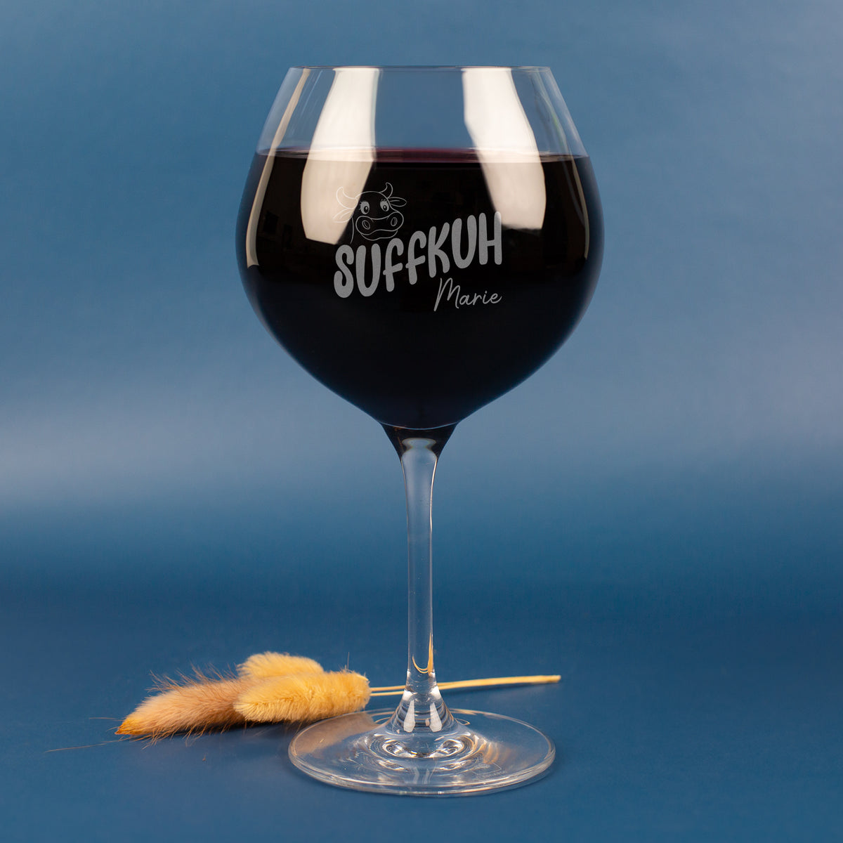 Personalisiertes Leonardo Weinglas mit Gravur „Suffkuh“ und Name Marie – witziges Burgunderglas aus klarem Glas, gefüllt mit Rotwein, vor blauem Hintergrund mit Deko-Halmen.