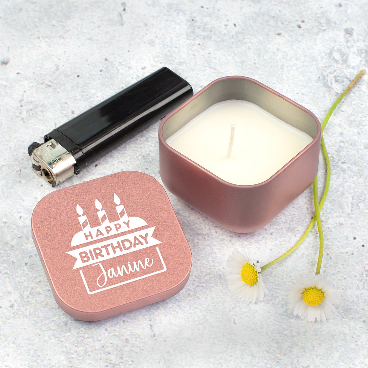 Roségoldene Mini Duftkerze mit Name Janine neben Feuerzeug und kleinen Blumen auf grauem Untergrund