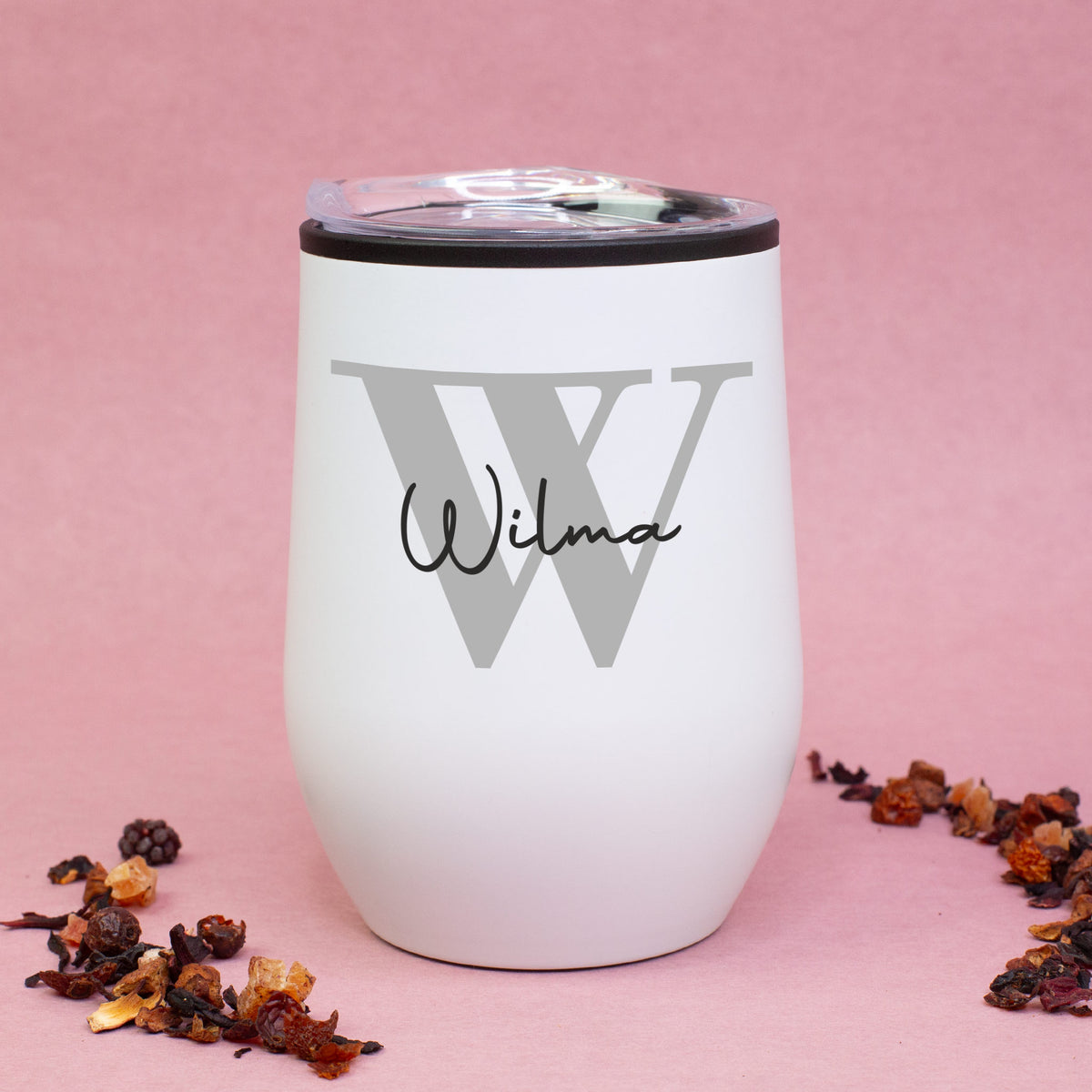 Weißer Thermobecher mit Initiale W und Name Wilma, auf rosa Hintergrund mit Teemischung dekoriert