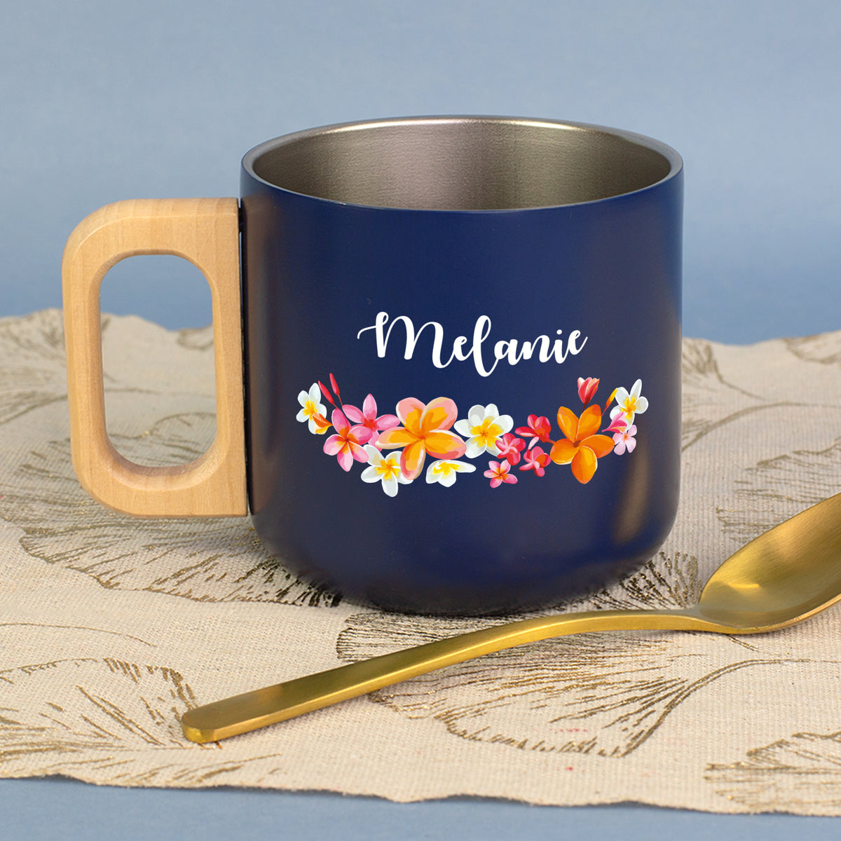 Dunkelblaue Metalltasse mit Holzgriff, Blumen-Motiv und Name Melanie neben goldfarbenem Löffel