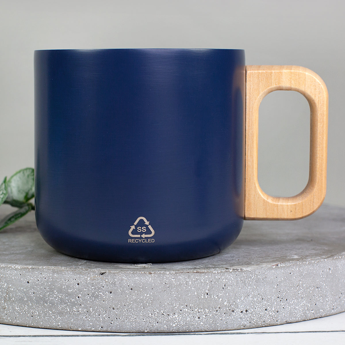 Rückseite der dunkelblauen Metalltasse mit Holzgriff und Recycled-Symbol auf Betonuntergrund