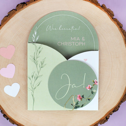 Geschlossene Hochzeitseinladung mit Ja Karte, floralen Details und goldener Klammer auf dekorativer Holzscheibe