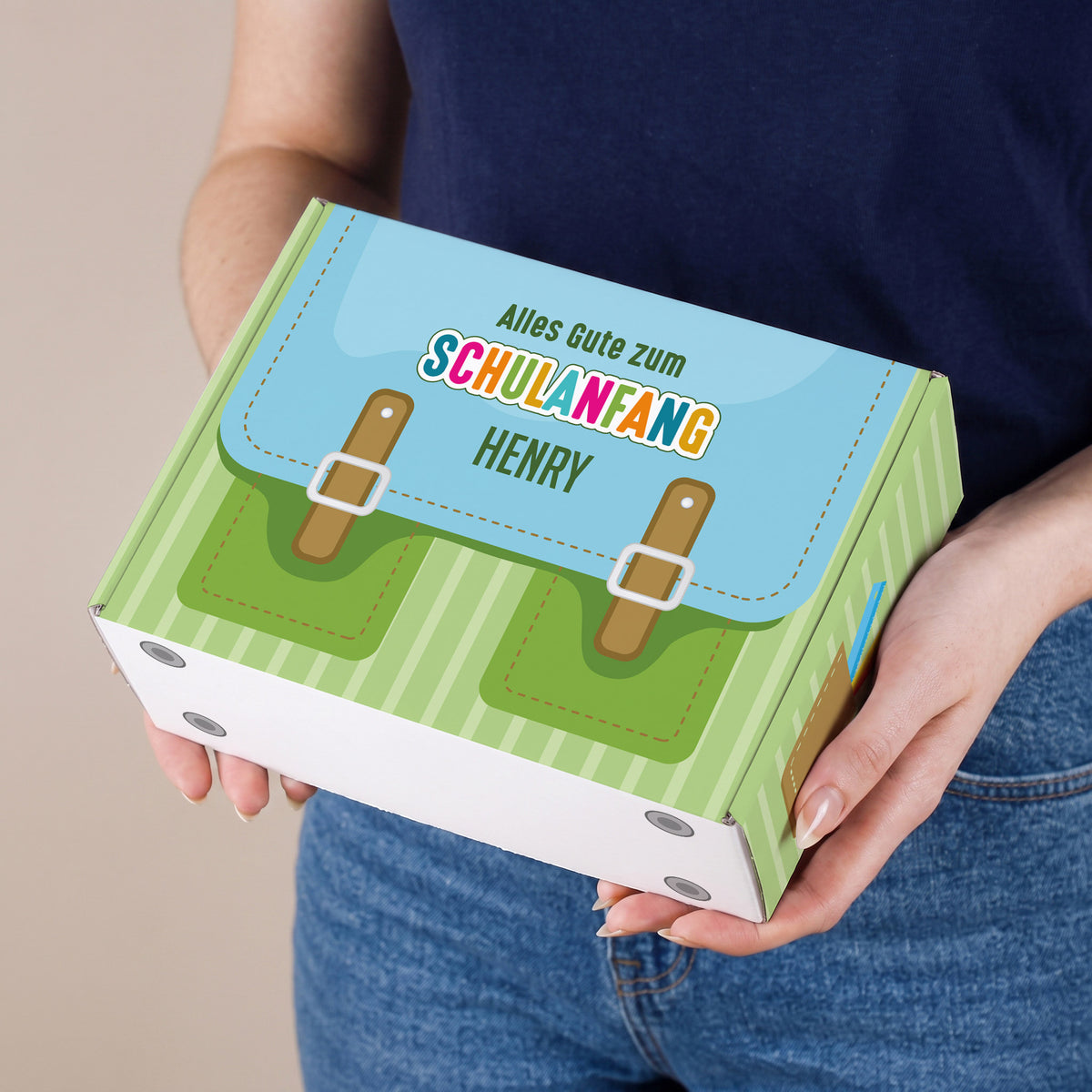 Kleine personalisierte Geschenkbox zur Einschulung in Grün mit Schulranzen-Design und Wunschname, von einer Person präsentiert