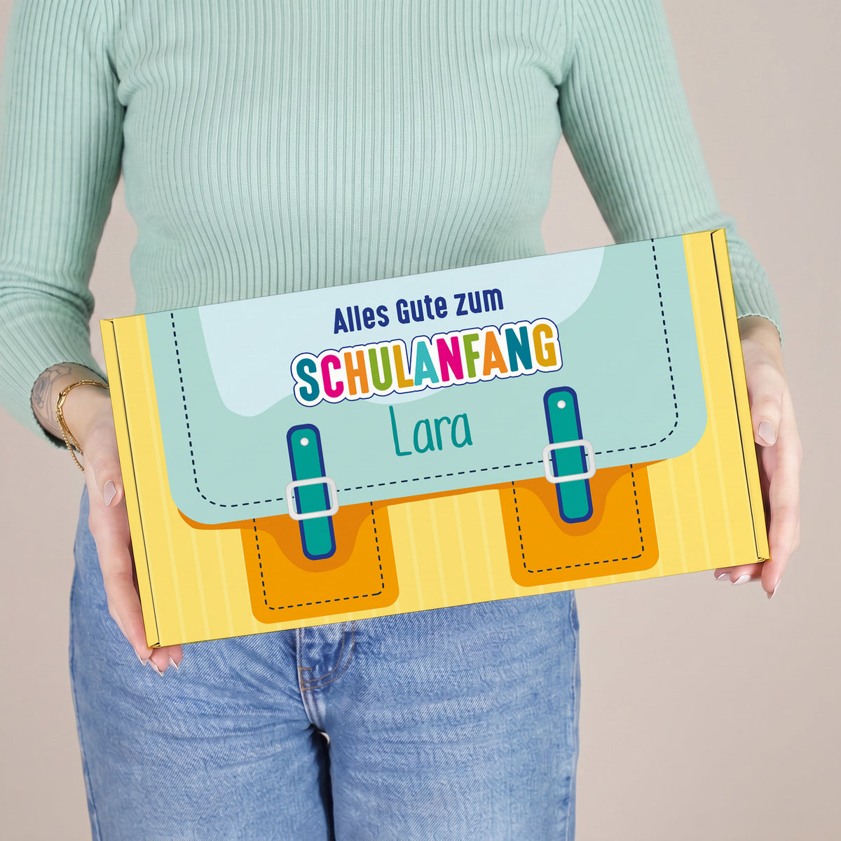 Mittlerer personalisierter Geschenkkarton zur Einschulung in gelb mit Schulranzen-Motiv und Name, von einer Person präsentiert