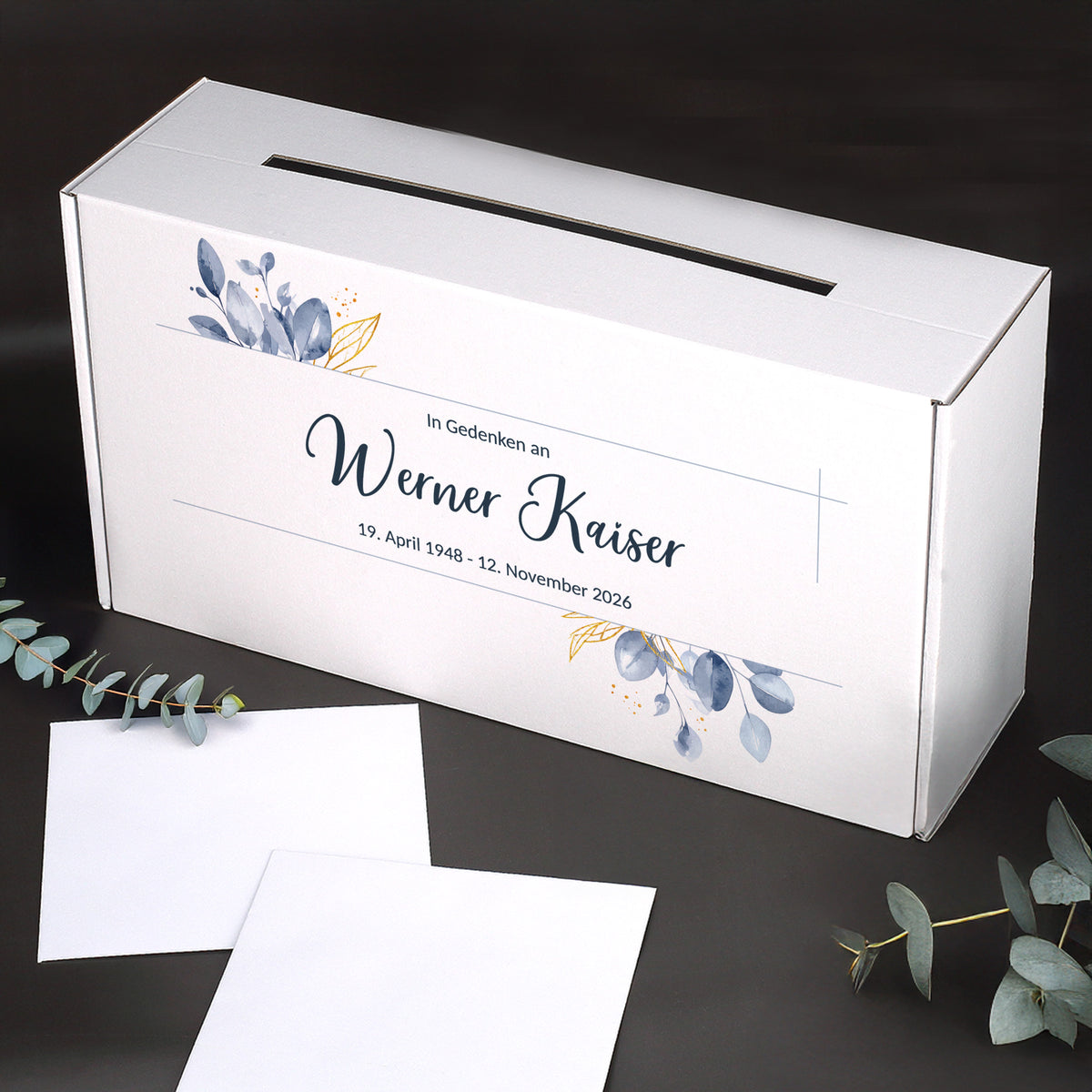 Personalisierte weiße Briefbox für Trauerfeiern mit Blattmotiv, Name und Datum, stabiler Karton, seitlicher Schlitz, dekoriert mit Eukalyptuszweigen.