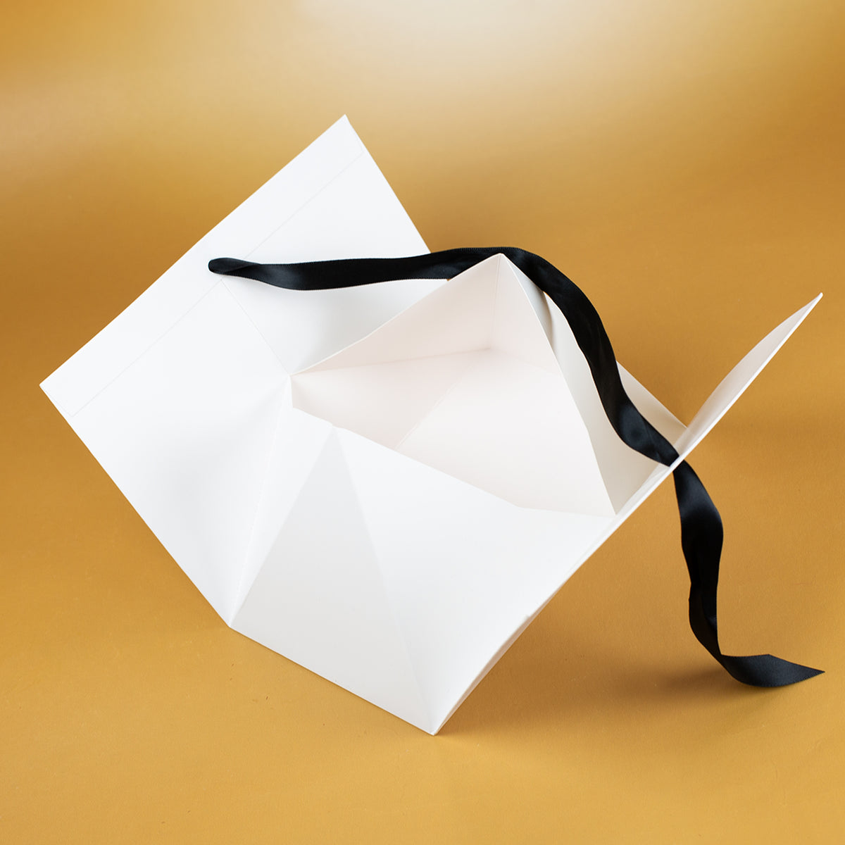 Innenansicht der geöffneten Geschenktasche aus weißem Karton, mit sichtbarer Faltkonstruktion und schwarzem Satinband, liegend auf goldenem Untergrund.