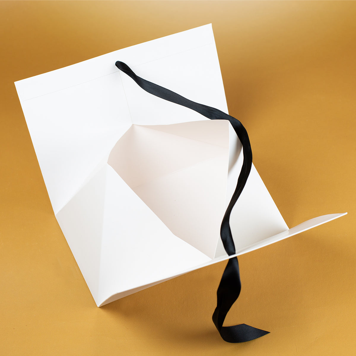 Innenansicht der geöffneten Geschenktasche aus weißem Karton, mit sichtbarer Faltkonstruktion und losem schwarzen Satinband, liegend auf goldenem Untergrund.