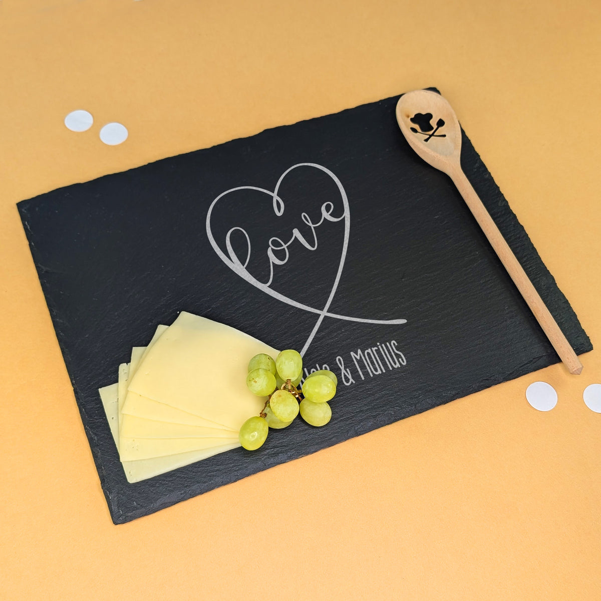 Schieferplatte mit Gravur „love“ in Herzform und „Nele & Marius“, dekoriert mit Käse, Trauben und Kochlöffel – auf beigem Hintergrund