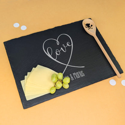 Schieferplatte mit Gravur „love“ in Herzform und „Nele & Marius“, dekoriert mit Käse, Trauben und Kochlöffel – auf beigem Hintergrund