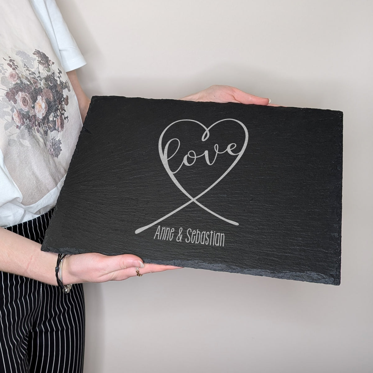 Schieferplatte mit Gravur „love“ in Herzform und „Anne & Sebastian“, von einer Person gehalten zur Größenansicht