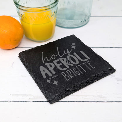 Untersetzer mit Gravur „holy Aperoli von Brigitte“ neben Saftglas und Orange