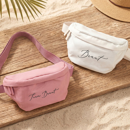 Rosa Bauchtasche mit Wunschtext Team Braut und cremefarbene Tasche mit Braut-Schriftzug auf Holzuntergrund