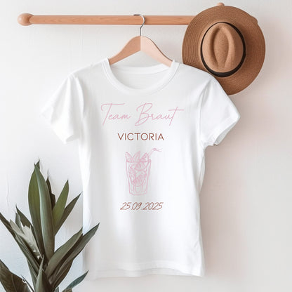Weißes T-Shirt mit Aufschrift „Team Braut“ in Rosé, darunter Name Victoria, Motiv mit Cocktailglas und Datum 25.09.2025.