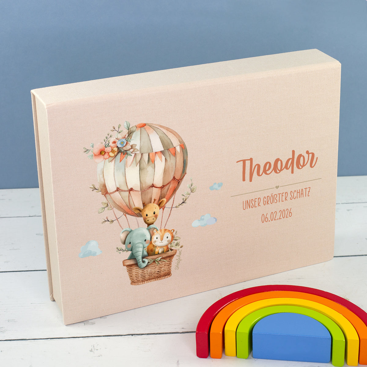 Beige Baby-Erinnerungsbox in Buchform mit Heißluftballon, Tiermotiv, Name und Wunschtext neben Regenbogen-Deko