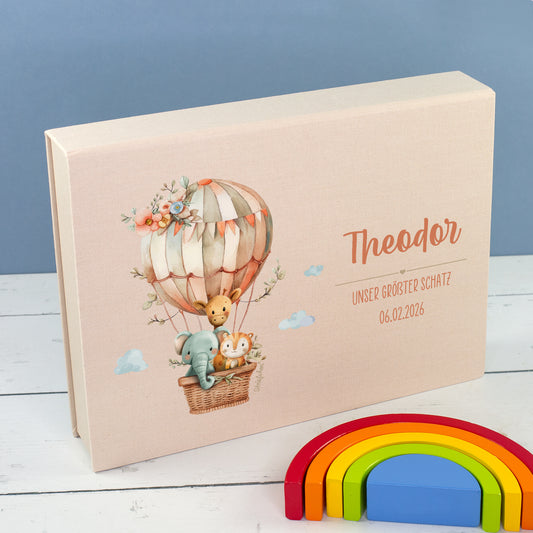 Beige Baby-Erinnerungsbox in Buchform mit Heißluftballon, Tiermotiv, Name und Wunschtext neben Regenbogen-Deko