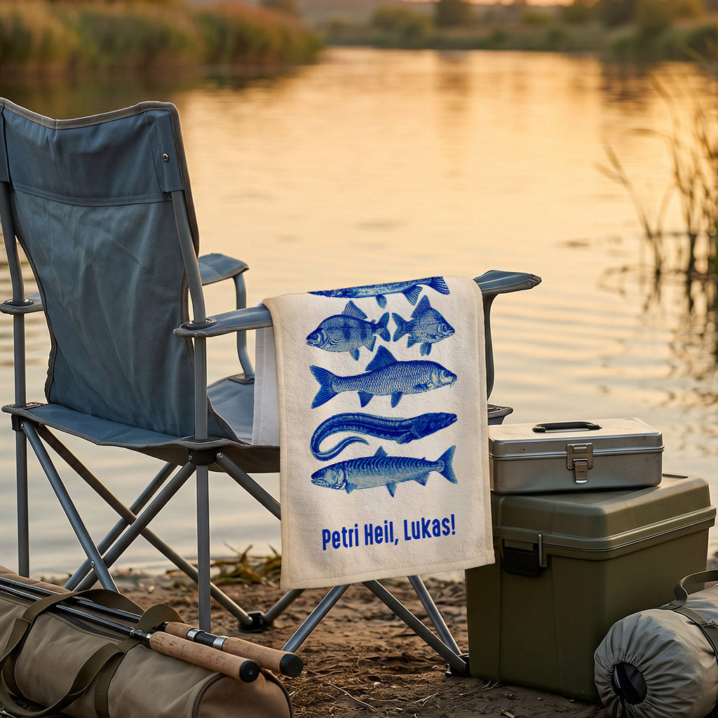 Personalisiertes Angler-Handtuch mit blauen Fischmotiven und Wunschtext auf einem Campingstuhl am See