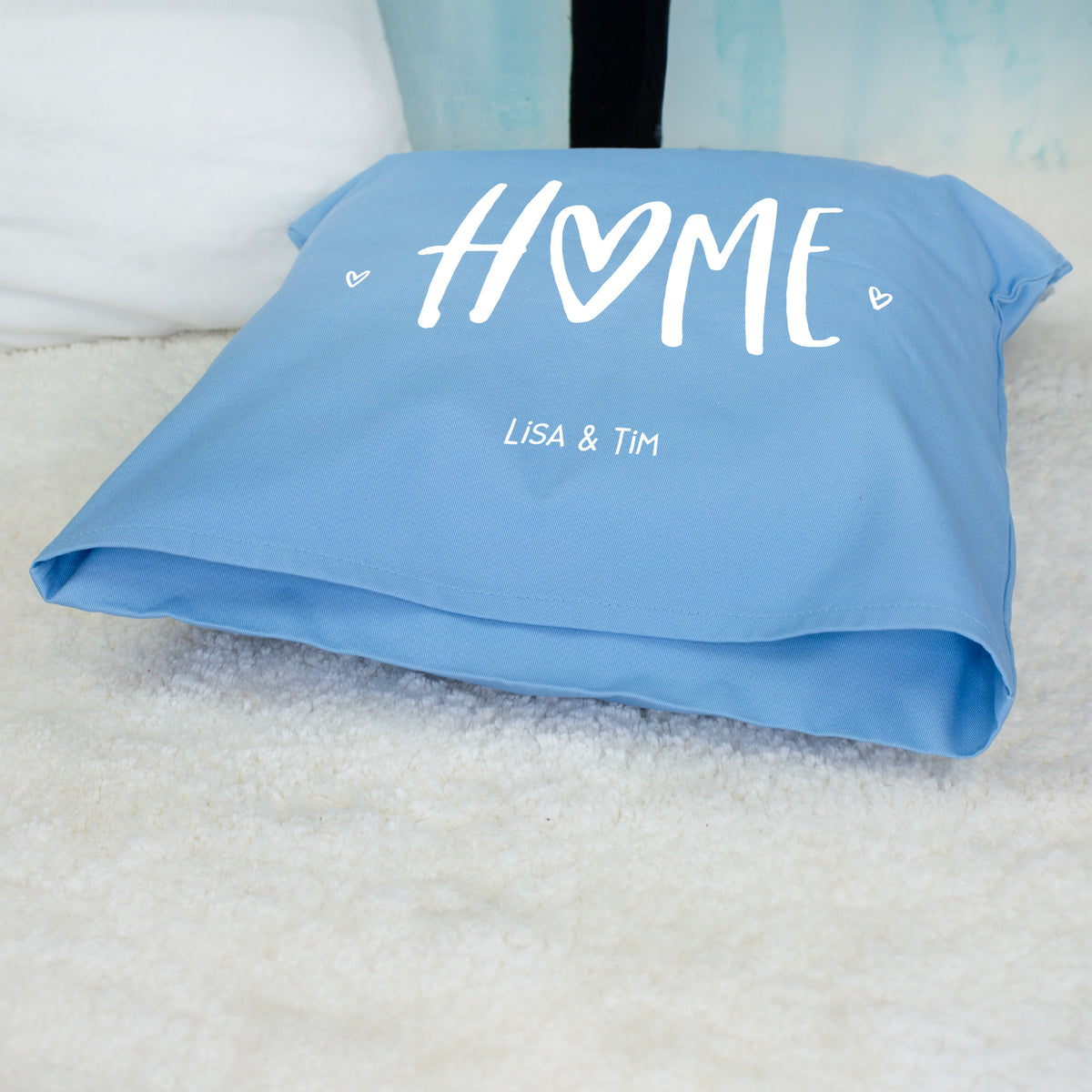 Blaues personalisiertes HOME Kissen mit Wunschtext Lisa und Tim liegend auf Fell