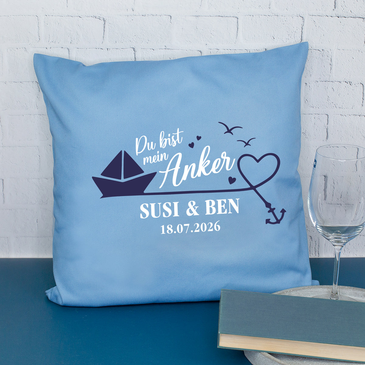 Personalisiertes Kissen in Blau mit Namen und Datum neben Glas und Buch