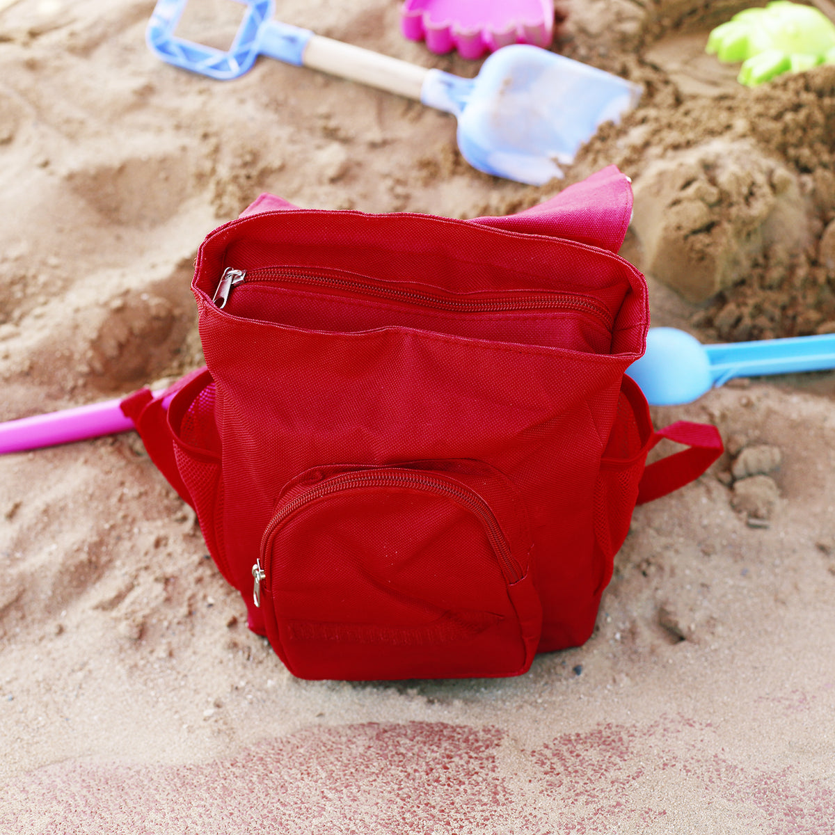 Geöffneter roter Kinderrucksack mit Hauptfach, Außentasche und seitlichem Netzfach auf dem Spielplatz