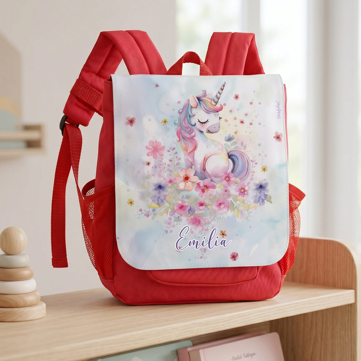 Personalisierter Kinder Rucksack in Rot mit Einhorn, Blumenmotiv und Name Emilia auf heller Holzbank