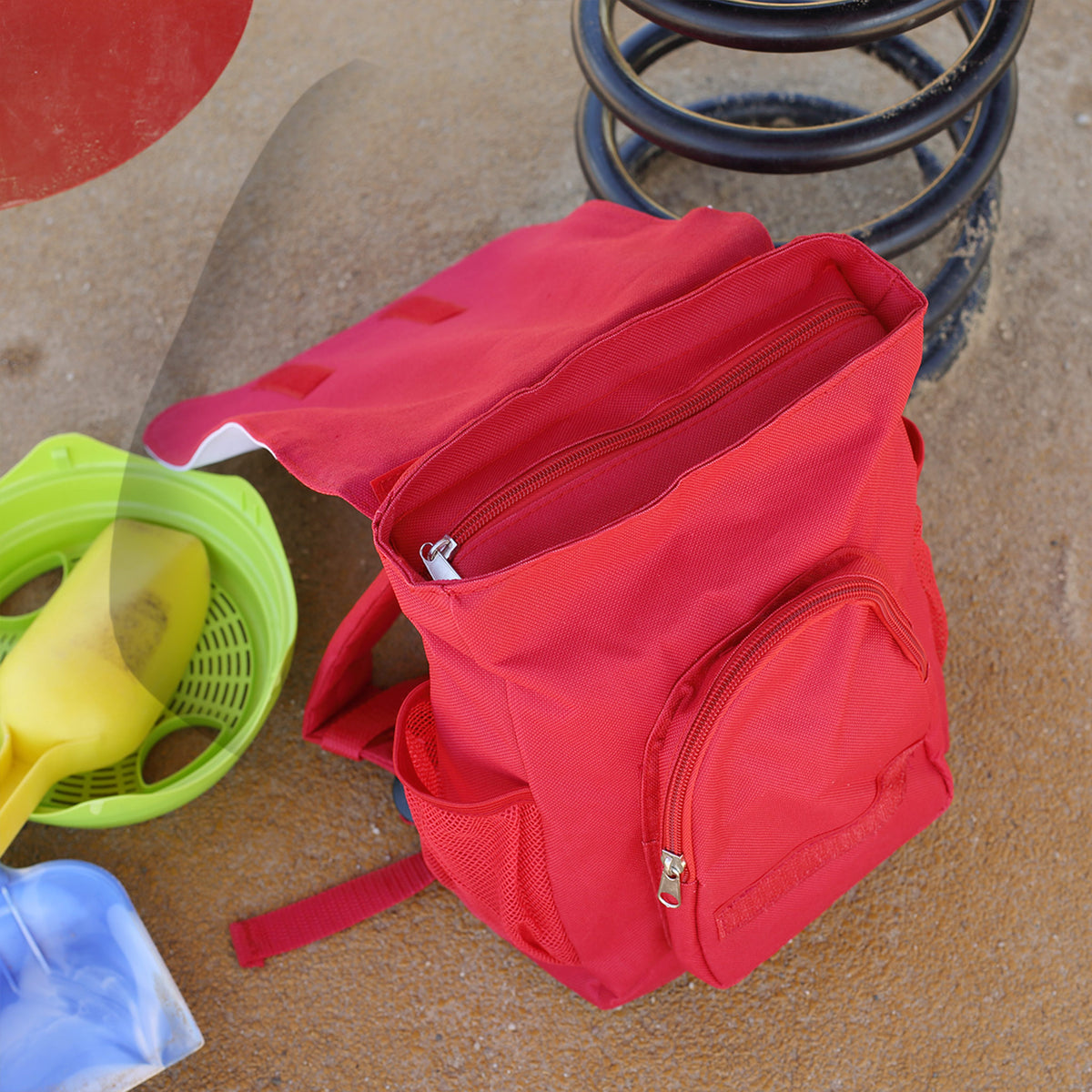 Geöffneter roter Kinderrucksack mit Hauptfach, Außentasche und Reißverschlüssen auf dem Spielplatz