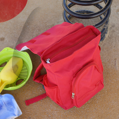Geöffneter roter Kinderrucksack mit Hauptfach, Außentasche und Reißverschlüssen auf dem Spielplatz