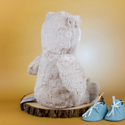 Rückansicht der flauschigen Eule, die auf einer Holzscheibe sitzt. Neben ihr liegen kleine blaue Babyschuhe – das Bild zeigt das weiche Fell und die handwerkliche Verarbeitung des Kuscheltiers.