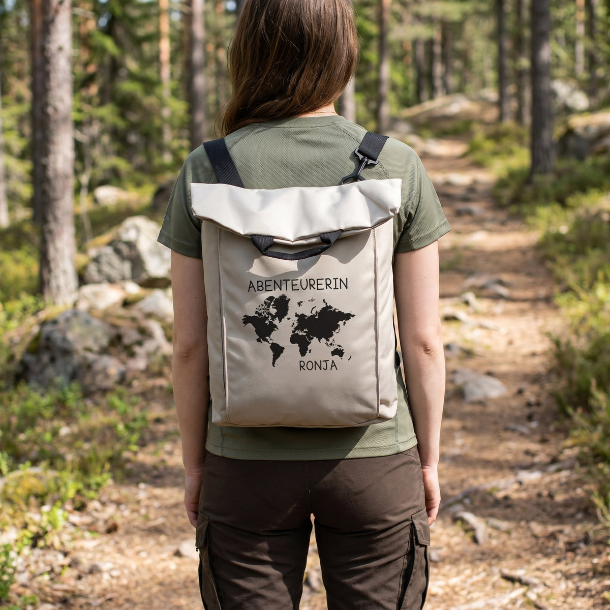 Frau trägt beigen personalisierten 2-in-1 Rucksack mit Weltkarten-Motiv, Wunschtext „Abenteurerin“ und Name „Ronja“ auf einem Waldweg