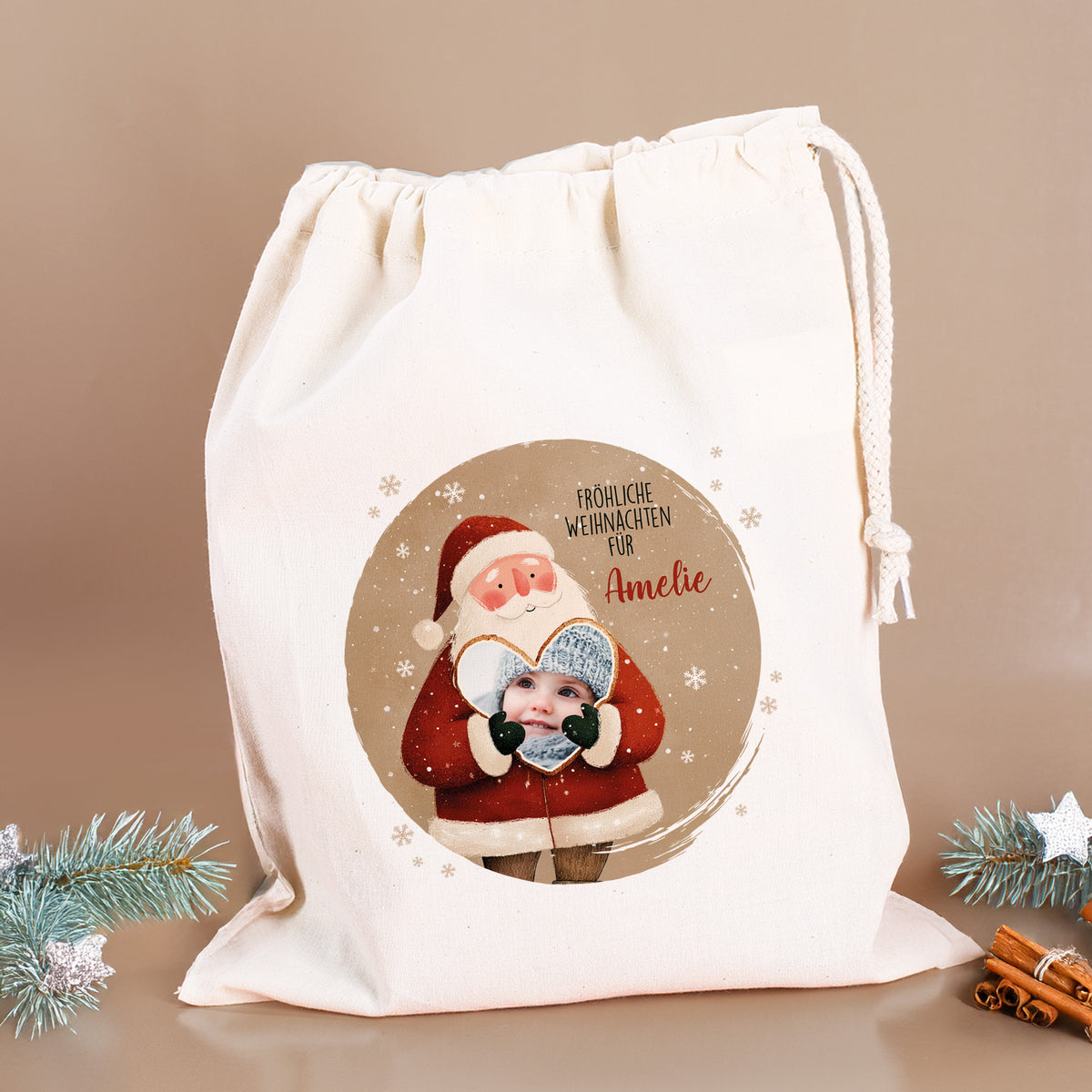 Großer Geschenksack mit Weihnachtsmann und Foto im Herz, Text „Fröhliche Weihnachten für Amelie“, auf beigem Hintergrund mit weihnachtlicher Deko aus Zweigen und Zimtstangen.