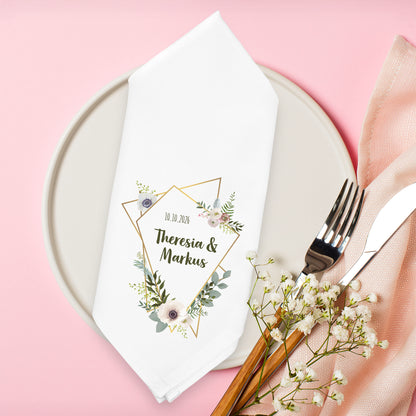Personalisierte Serviette mit floralen Blüten, Eukalyptus und geometrischem Goldrahmen auf gedecktem Tisch