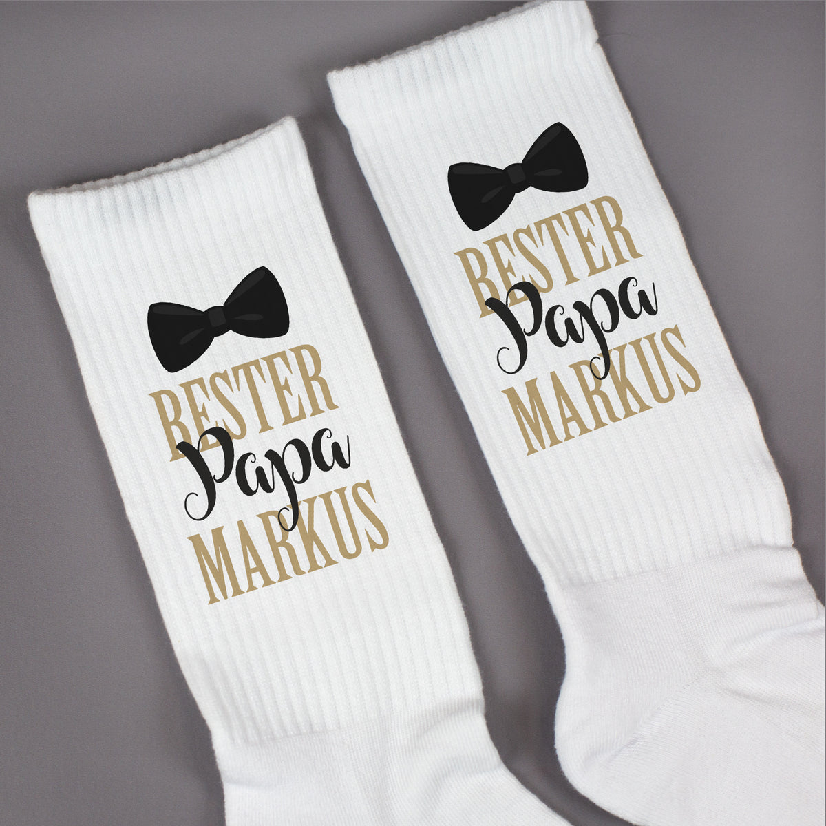 Paar weiße Socken mit „Bester Papa“-Motiv und Name auf grauem Hintergrund