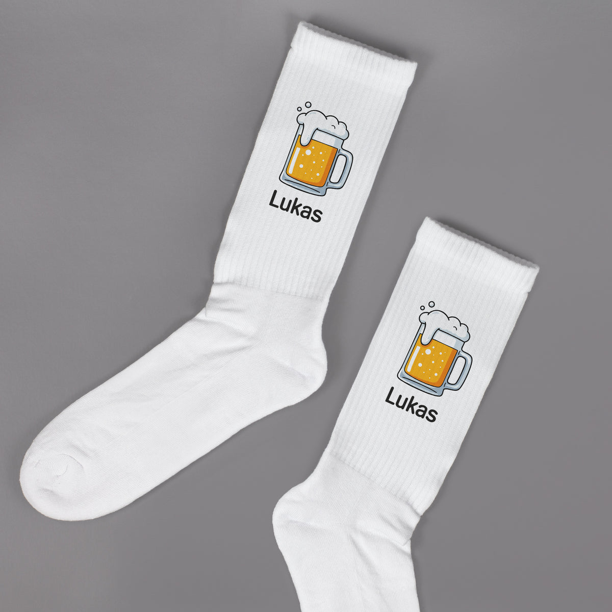 Paar personalisierte Socken mit Bierkrug-Motiv und Name „Lukas“ – flachliegend auf grauem Hintergrund.