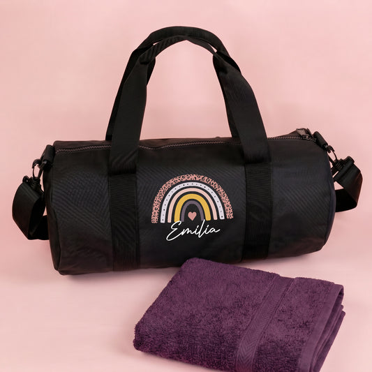 Personalisierte schwarze Sporttasche mit Regenbogen Motiv und Name Emilia neben lila Handtuch