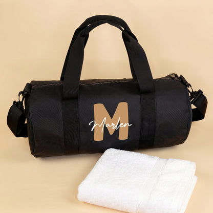 Schwarze Sporttasche mit großer brauner Initiale M und weißem Namen Marlen vor hellem Hintergrund mit Handtuch
