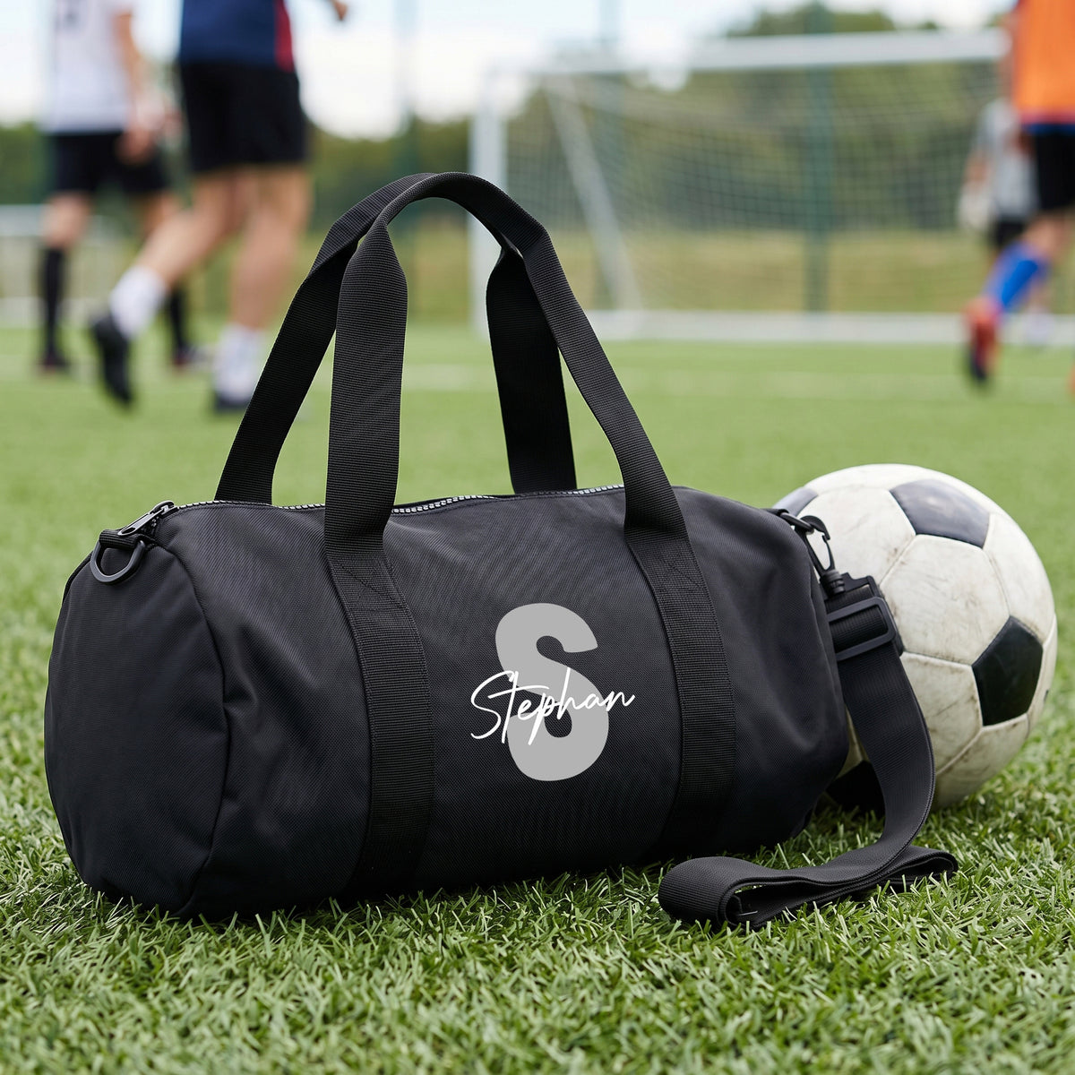 Personalisierte schwarze Sporttasche mit grauer Initiale S und Name Stephan auf Fußballplatz neben einem Fußball