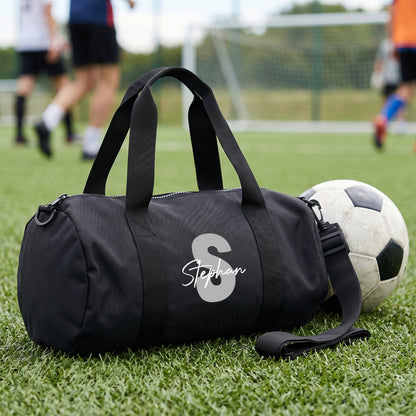 Personalisierte schwarze Sporttasche mit grauer Initiale S und Name Stephan auf Fußballplatz neben einem Fußball