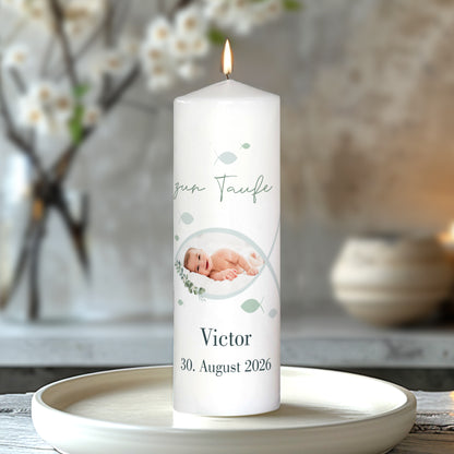 Nahaufnahme einer personalisierten Taufkerze mit Fischsymbol und integriertem Foto. Der Schriftzug „zur Taufe“, das Foto des Babys sowie Name „Victor“ und Datum „30. August 2026“ sind gut lesbar. Die Kerze steht brennend auf einem hellen Teller, dekoriert mit Blüten.